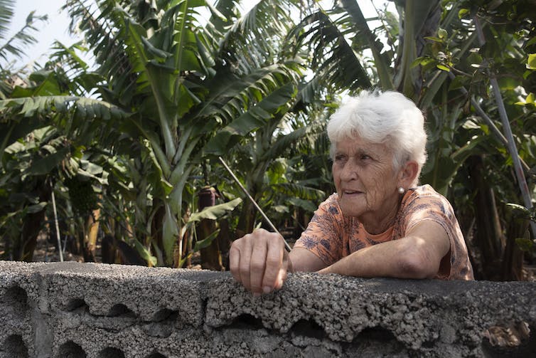 Una mujer madura se apoya contra un muro de piedra con plátanos al fondo.
