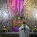 A priest wearing a face mask and white vestments holds a microphone and stands in front of a stone wall with religious carvings and purple light