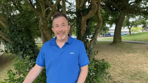 Chishdam Colin Weston con un polo azul frente a un árbol cubierto de hiedra, Stratford Park en Stroud. El estacionamiento y la carretera peatonal son visibles en el fondo. 