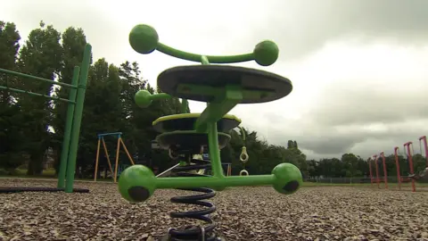 Una pieza de equipo de BBC Play Park, un jinete de primavera, es verde brillante y tiene dos asientos. El resto del parque se puede ver en el fondo, con nubes oscuras sobre la cabeza. 