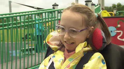 Foto de la hija de Sophie Millie. Está en un cochecito y usa un impermeable amarillo con vidrio rosa púrpura. Ella tiene cabello claro. 