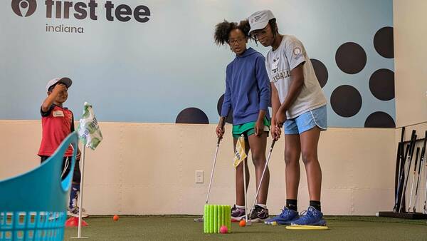 Tres niños practican el césped interno en el primer lugar de Tee Indiana. Un niño con una camisa roja mira a las dos chicas mayores con una camisa azul y gris combinan con su espuma.