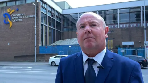 Jim O'Boyle, miembro del Consejo de la BBC, para el antiguo sitio web de CoV Baths. Lleva un traje azul y una corbata con una camisa blanca y se encuentra al otro lado del camino desde el edificio donde puede ver un letrero que dice el Centro de Deportes y Leisure de Coventry.