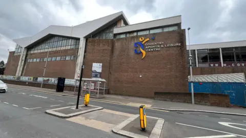 Vista del centro de deportes y ocio de Coventry de Coventry en Fairfax Street. El camino corre frente a un edificio de ladrillo con grandes ventanas de vidrio en la parte superior y en un techo desigual. El letrero que identifica el edificio como un