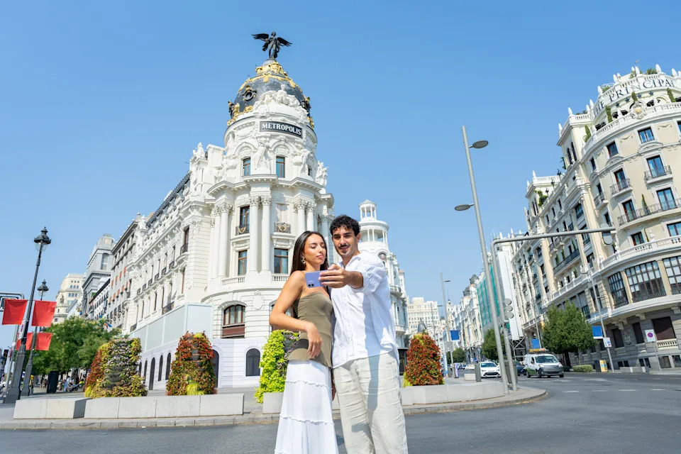 Pareja joven de vacaciones sonriendo sosteniendo un teléfono inteligente y tomándose un selfie frente al histórico edificio Metrópolis en Madrid, compartiendo experiencias de viaje. Una joven pareja se autorretrato frente al famoso edificio Metrópolis de la Gran Vía, plasmando un feliz recuerdo de viaje durante sus vacaciones. Un día soleado y una elegante arquitectura europea crean el escenario perfecto para el turismo moderno y el romance. Esta imagen representa el amor, la conexión y la aventura, ideal para ilustrar conceptos de estilo de vida digital, fotografía de viajes, recorridos por la ciudad y parejas disfrutando juntas de una escapada a la ciudad. Madrid, España