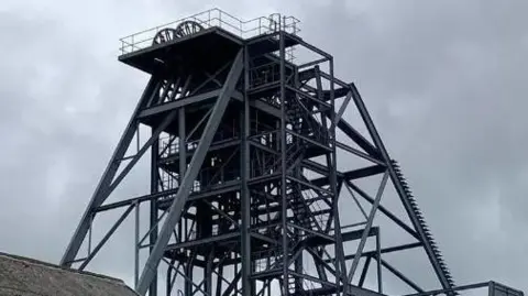 Mina de estaño de South Crofty. Una estructura de metal gris está suspendida sobre el suelo con una escalera/escalón que conduce a la cima. El cielo también está gris.