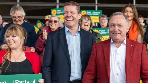 Getty Images Un grupo de personas con carteles de Plaid Cymru rodean al líder del partido Rhun ap Iorwerth, un hombre con traje azul marino y camisa azul, todos sonriendo.