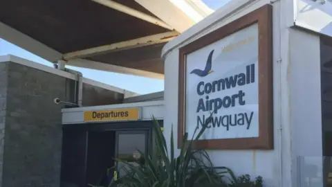 Entrada al aeropuerto de Cornwall Newquay con cartel en la pared curva. Las puertas automáticas son visibles con un cartel amarillo en la parte superior que dice 