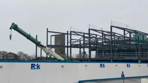 Una grúa, junto a una estructura de vigas de acero, detrás de un estante azul y blanco con la etiqueta Rok Construction.
