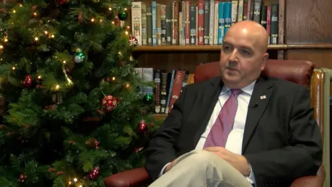 Jonathan Le Tocq, vestido con chaqueta oscura, camisa blanca y corbata rosa, está sentado con las piernas cruzadas en un sillón de cuero rojo junto a un árbol de Navidad decorado.