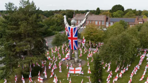 PA Media Una gran estatua plateada de un minero en Brownhills está adornada con una bandera Union Jack usada como capa con banderas cruzadas de San Jorge extendidas en el suelo donde se encuentra la estatua.