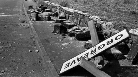 Foto en blanco y negro de PA Media de 1984. Señal de tráfico de la ciudad de Orgreave en el suelo, entre un montón de escombros. Junto a la carretera han derribado un muro y sobre las piedras del muro roto hay un cartel.