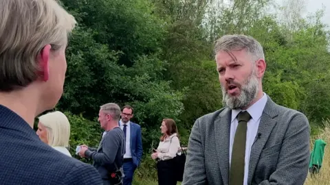 El editor político de la BBC de Yorkshire, James Vincent, entrevista a la diputada Yvette Cooper en el lugar de la llamada Batalla de Orgreave. Tiene una barba canosa y viste un traje a cuadros gris con una corbata verde. La Sra. Cooper está inclinada hacia la cámara y lleva un traje azul.