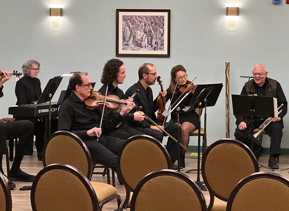 Músicos vestidos actúan en un pequeño conjunto con violines, trompeta y teclado en una sala con obras de arte enmarcadas en la pared.