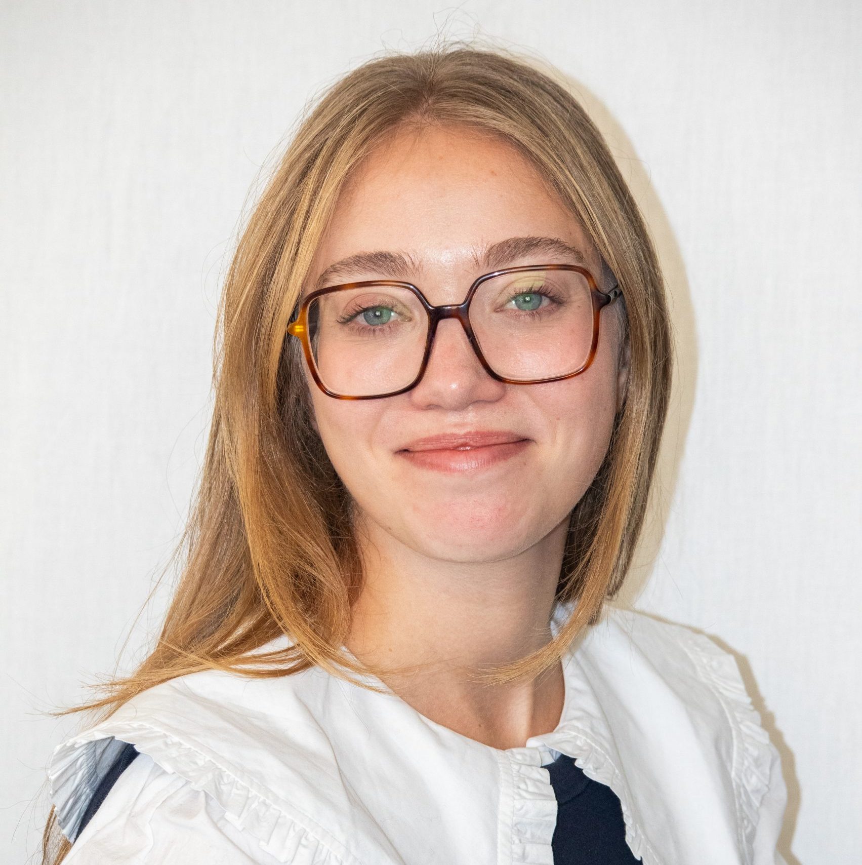 Una mujer de largo cabello rubio y gafas grandes, vestida con un top blanco con volantes, sonríe suavemente sobre un fondo claro.