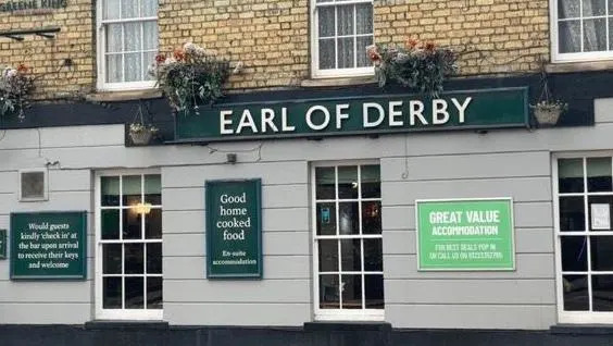 Pub Earl of Derby: edificio marrón y gris con marcos blancos. Palabras "Conde de Derby" están escritos en letras mayúsculas blancas en un letrero verde pegado al frente del pub. Debajo del cartel hay carteles publicitarios. "buena comida casera" y "alojamiento de gran valor".
