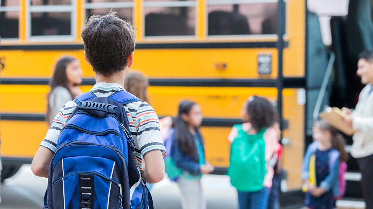 Vista trasera de un niño de primaria esperando abordar un autobús escolar. Sus compañeros de clase suben al autobús al fondo.