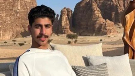 Mohammed Algasim lleva una camisa blanca con una franja azul sobre los hombros. Está sentado en un sofá con almohadones color crema a su lado. Detrás parece haber una escena desértica con una superficie de arena/roca y estructuras rocosas altas.