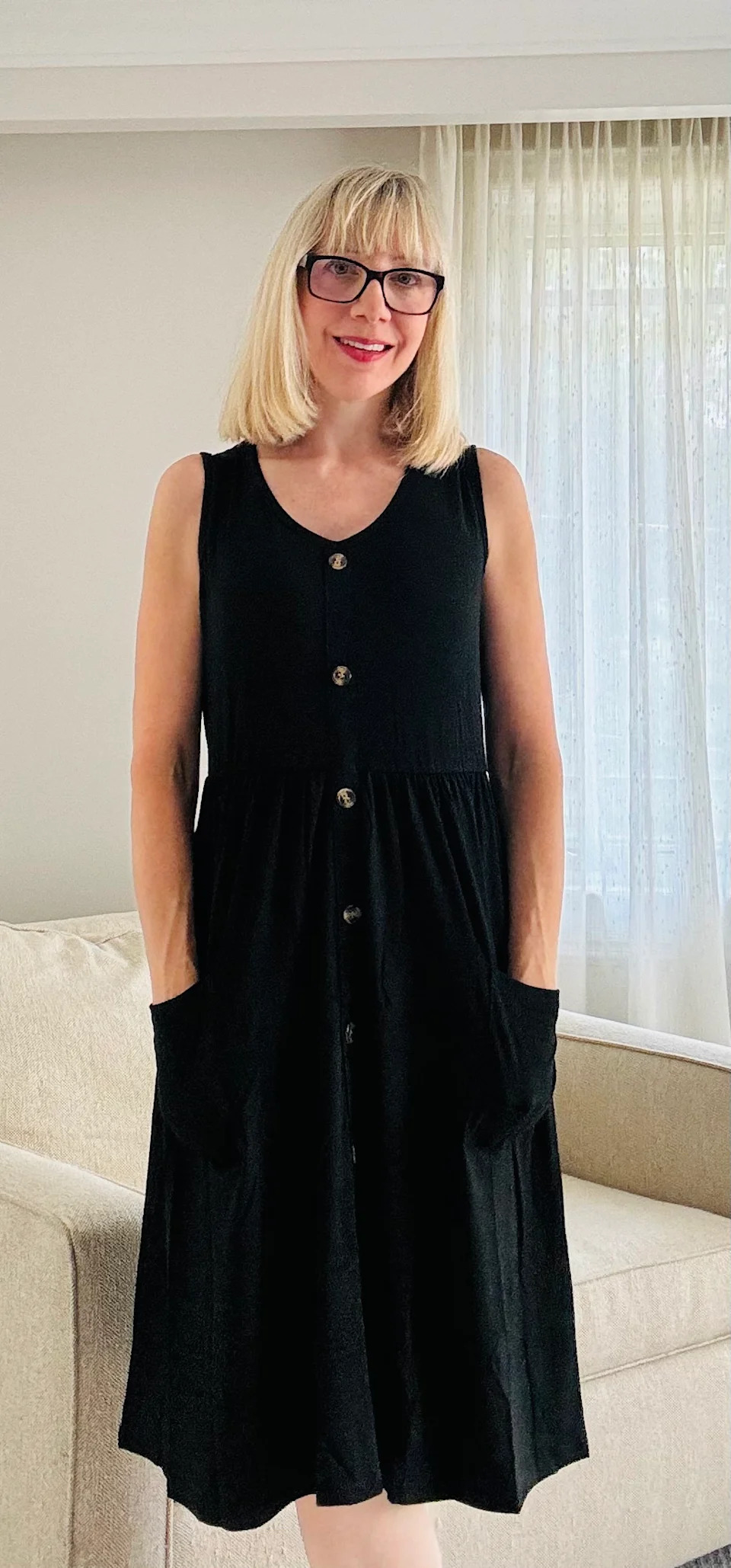 Mujer rubia con gafas, vestida con un vestido negro con botones.