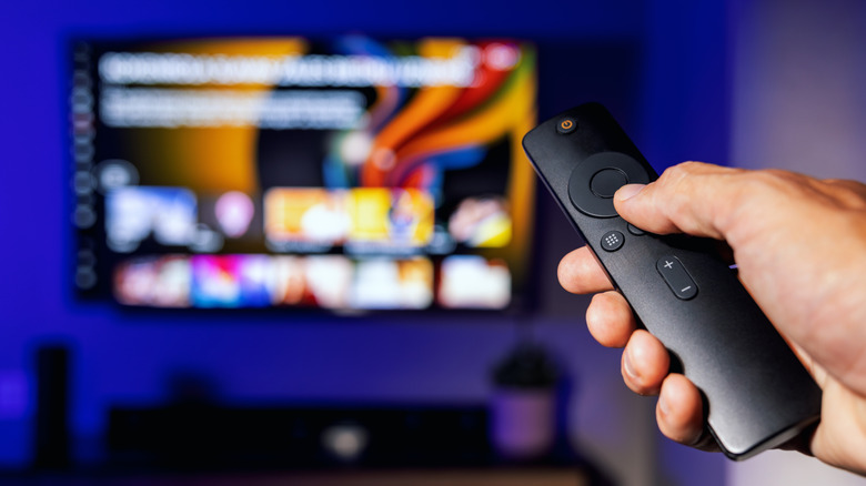 Alguien está usando el control remoto de un televisor con un televisor inteligente de fondo.